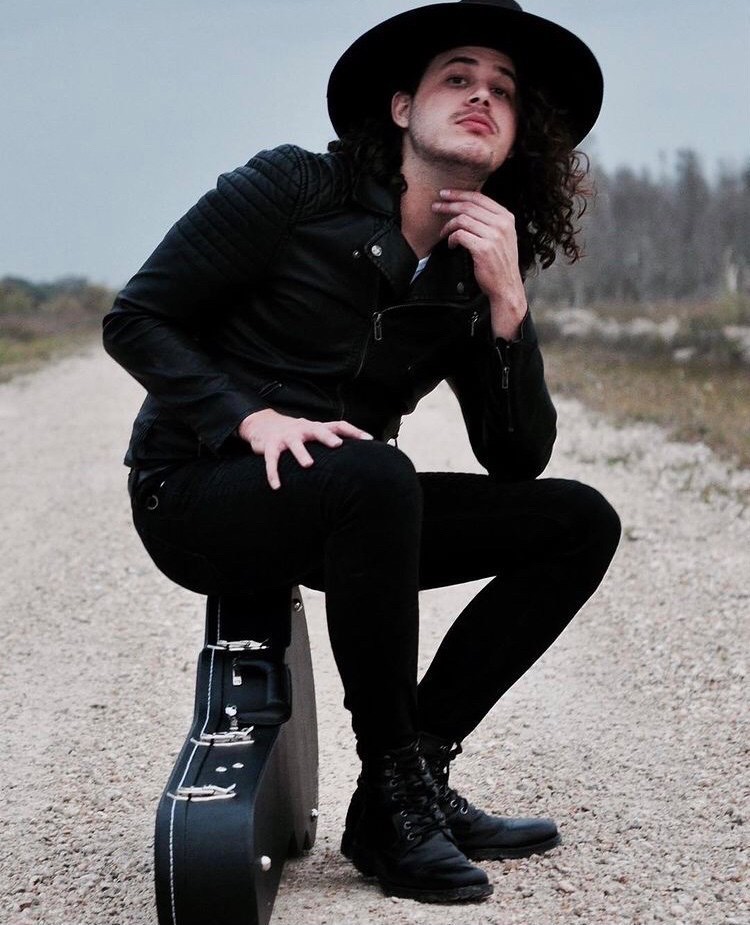 Outdoor artist portrait of Will Plus seated on a guitar case in a black hat and leather jacket