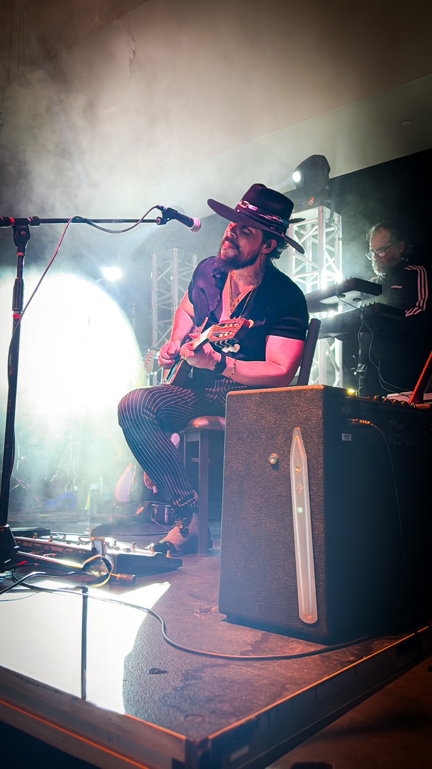Will Plus seated on stage playing electric guitar beneath dramatic concert lights and smoke